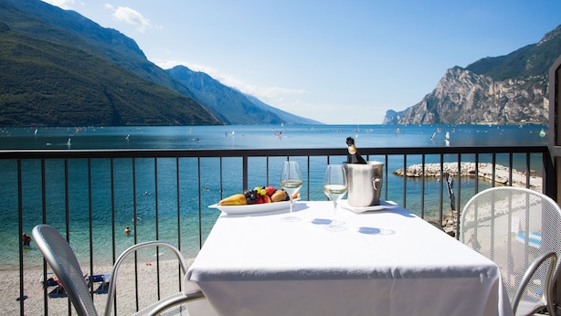 Una mesa con mantel blanco, una botella de champán y un plato de fruta en un balcón con vistas a un lago y montañas al fondo.