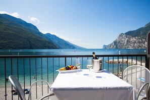 Een tafel met een witte tafelkleed, een fles champagne en een fruitschaal op een balkon met uitzicht op een meer en bergen op de achtergrond.