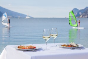 Ein Tisch mit zwei Tellern Essen und zwei Gläsern Weißwein, vor einer ruhigen See mit entfernten Segelbooten.