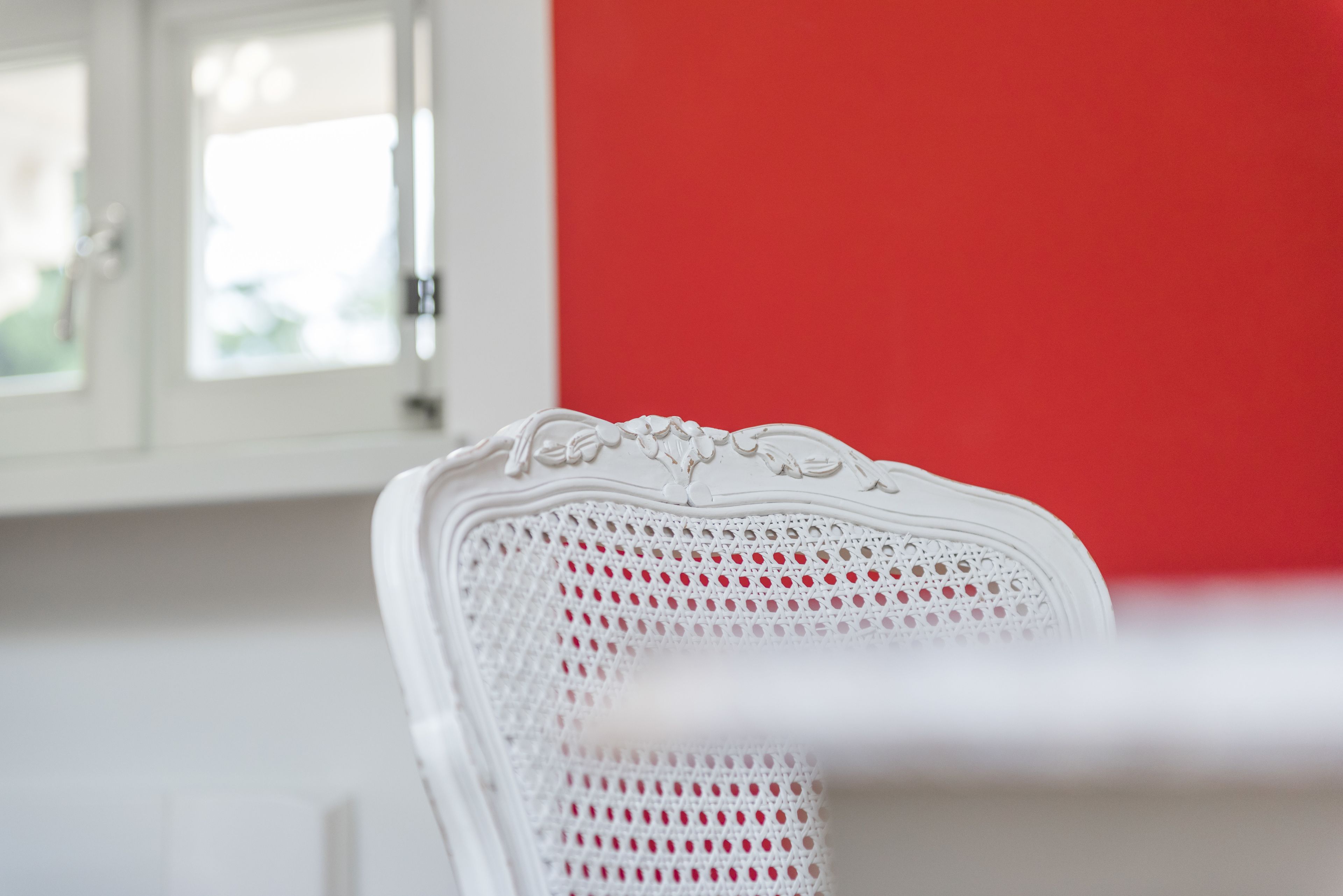 Una silla blanca con respaldo perforado y detalles ornamentales, ubicada en una habitación con pared roja y ventana con vistas a la vegetación.