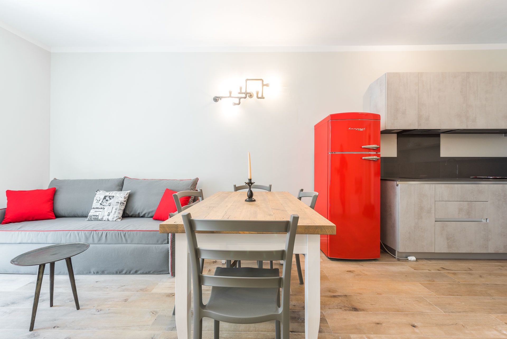 Un espace de vie compact comprenant une table à manger, un réfrigérateur rouge, un canapé gris avec des coussins rouges et une petite table d’appoint.