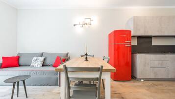 Un espace de vie compact comprenant une table à manger, un réfrigérateur rouge, un canapé gris avec des coussins rouges et une petite table d’appoint.