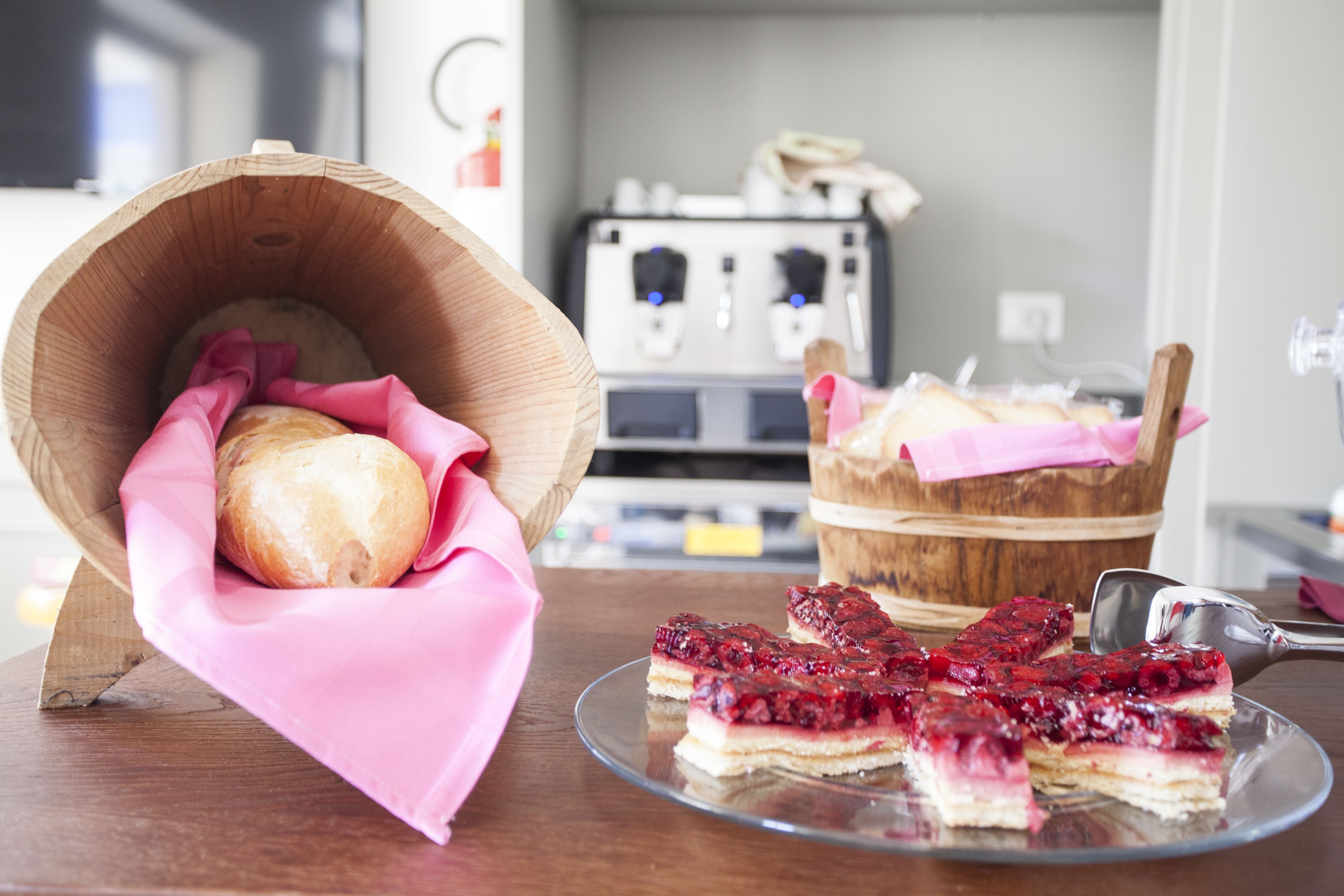 Una cesta de madera con un pan, un paño rosa y un cubo de madera con pan, además de un plato de vidrio con porciones de tarta de frutos rojos.