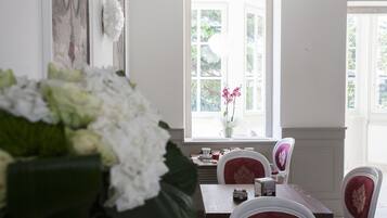 A dining area with a table set for four, white chairs with red cushions, a vase with flowers, and a view through a window with white shutters.
