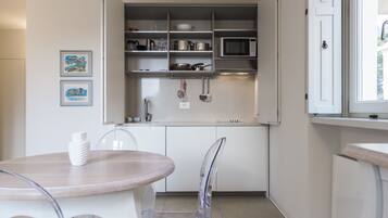Cocina compacta con alacenas blancas, microondas y varios utensilios. Hay una mesa de comedor con sillas y dos cuadros en la pared.