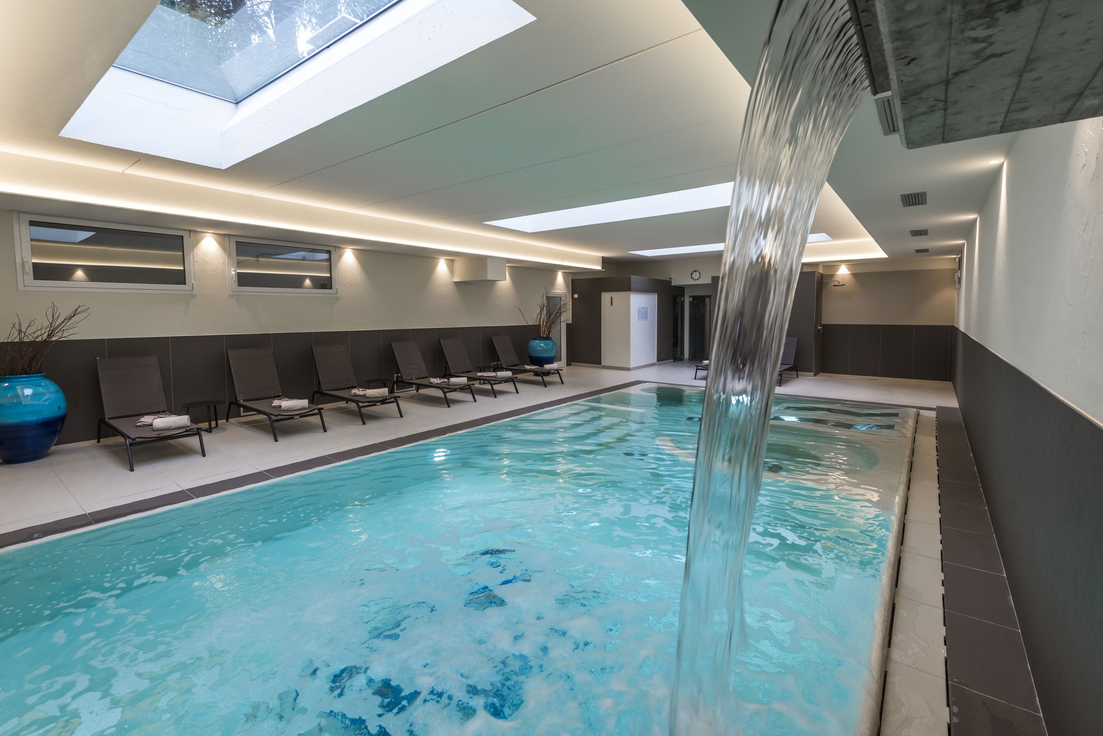A modern indoor swimming pool with a skylight, lounge chairs, and a water feature.