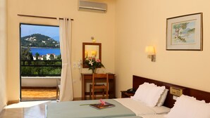 A hotel room with a bed, a desk with a chair, a small table with a vase of flowers, a framed picture on the wall, and a view of a lake and buildings through a window. - Paradise Hotel Corfu (Corfu)