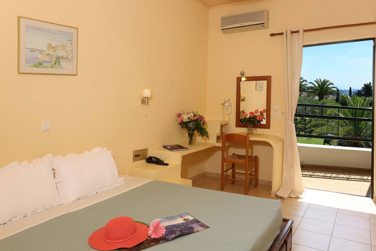 A hotel room with a bed, a desk with a chair, a bathroom mirror, and a balcony with a view of greenery.
