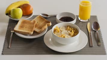 Desayuno con tostadas, fruta, un tazón de cereal, mermelada y jugo de naranja sobre una mesa.