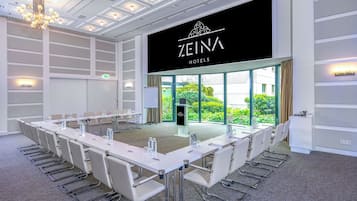 A conference room with a long table, chairs, and a chandelier. The room has a large window with a view of greenery outside.