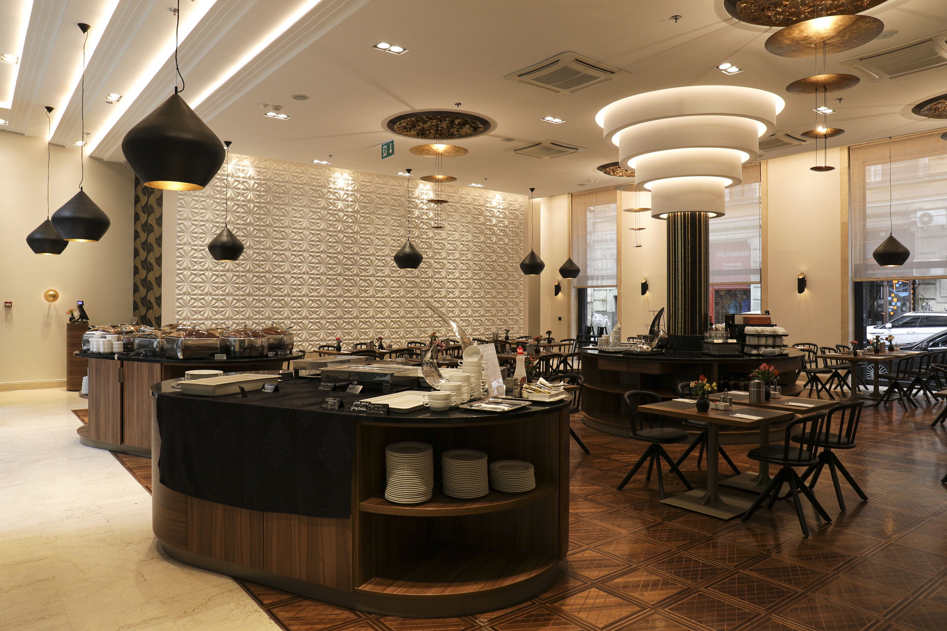 A modern restaurant interior with a central counter, multiple dining tables, and hanging pendant lights.