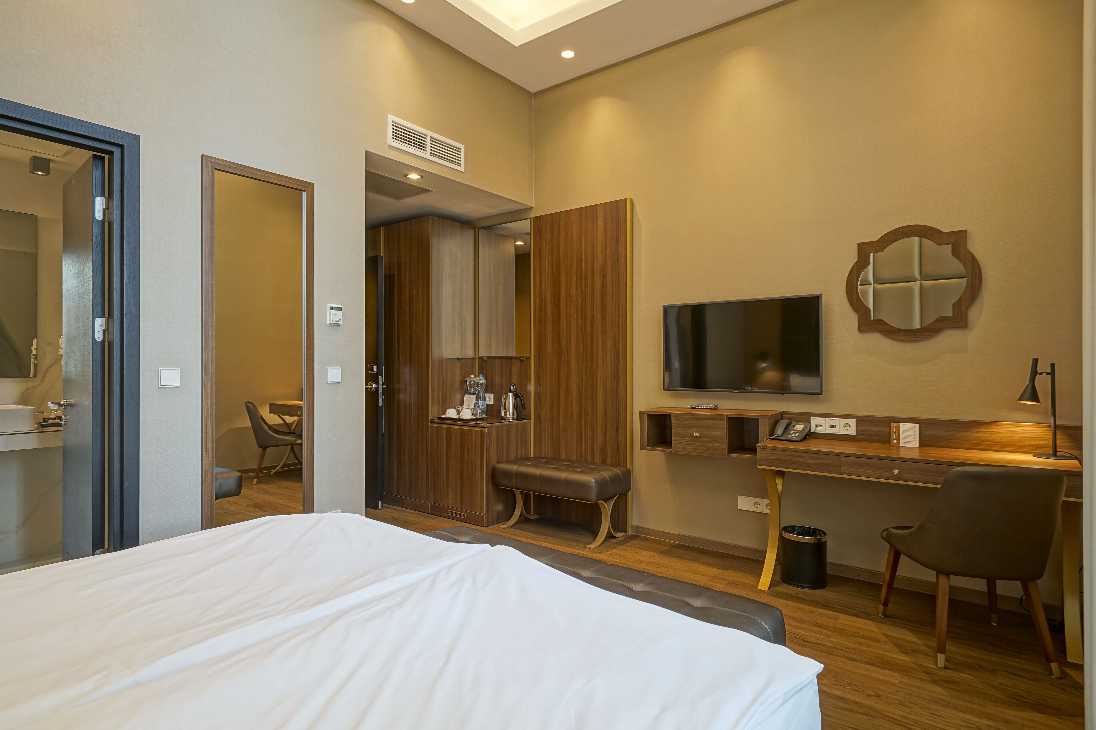 A hotel room with a bed, a desk with a computer, a TV, a mirror, and a small seating area.