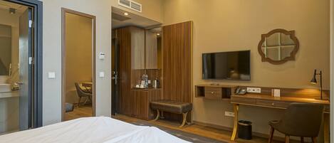 Habitación de hotel con cama, escritorio con computadora, televisor, espejo y un pequeño rincón de descanso.