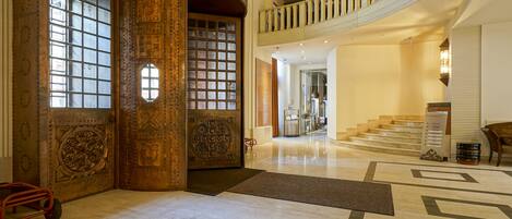 Un lobby imponente con puertas decoradas, piso de mármol y una escalera que conduce a un nivel superior.
