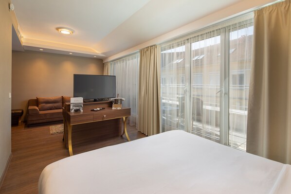 A hotel room with a large bed, a wooden desk with a flat-screen TV, a sofa, and a large window with curtains.