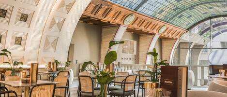 Un espacio interior amplio con techos altos, una cúpula de cristal y mobiliario moderno, incluyendo mesas redondas y sillas de mimbre.