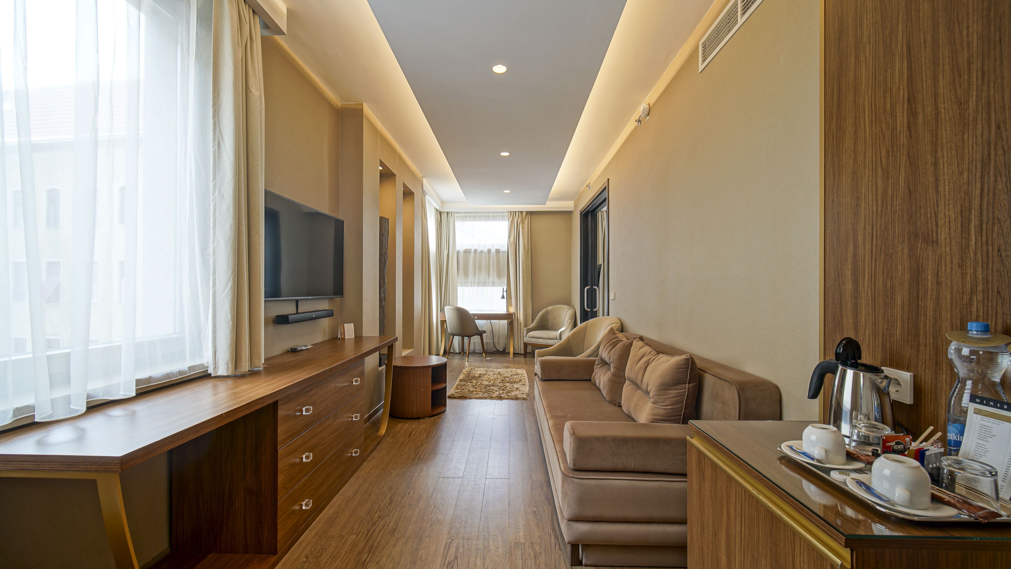 Een moderne hotelkamer met een houten vloer, een flatscreen-tv, een bank, een eettafel met een dienblad vol bekers en een waterkoker, en een groot raam met dunne gordijnen.
