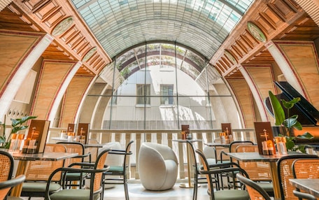 A spacious indoor area with a high, arched glass ceiling and a central seating area with modern chairs and tables.