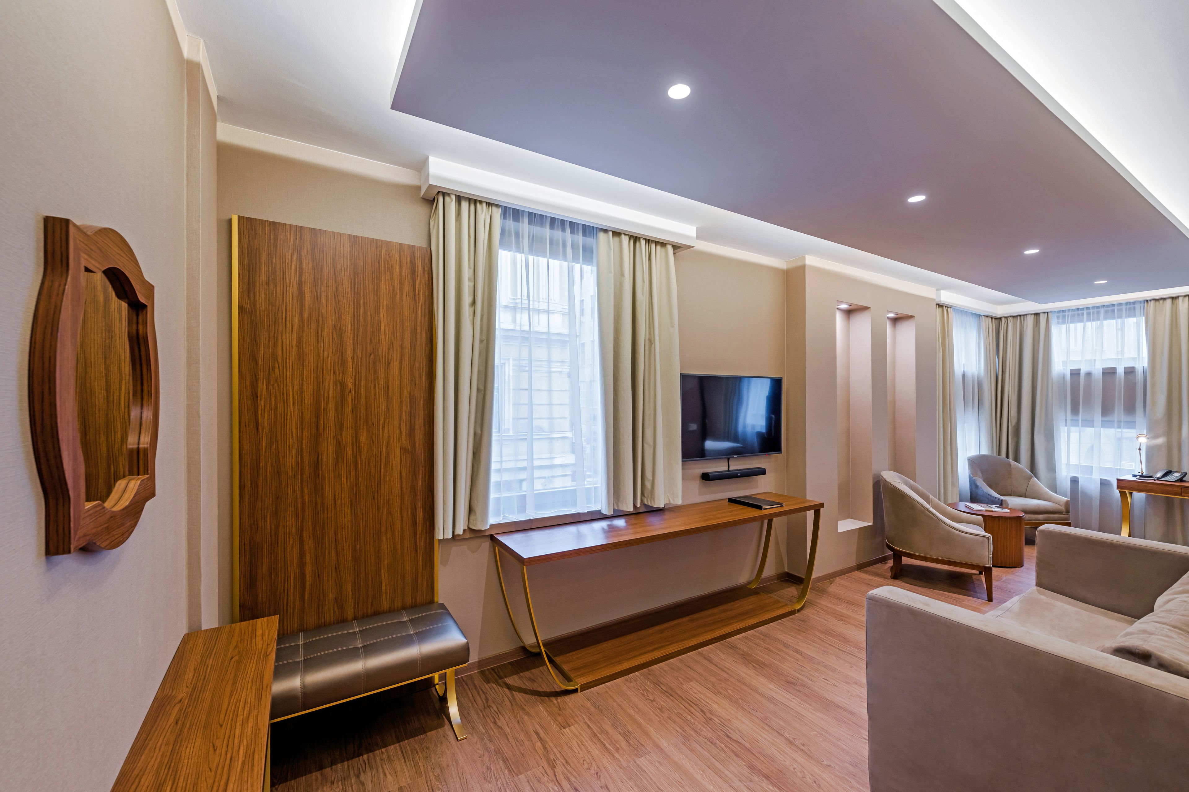 A modern hotel room with a wooden bench, a sofa, a TV, and a desk.