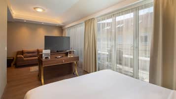 A hotel room with a large bed, a flat-screen TV, a wooden desk, a sofa, and a balcony with curtains.