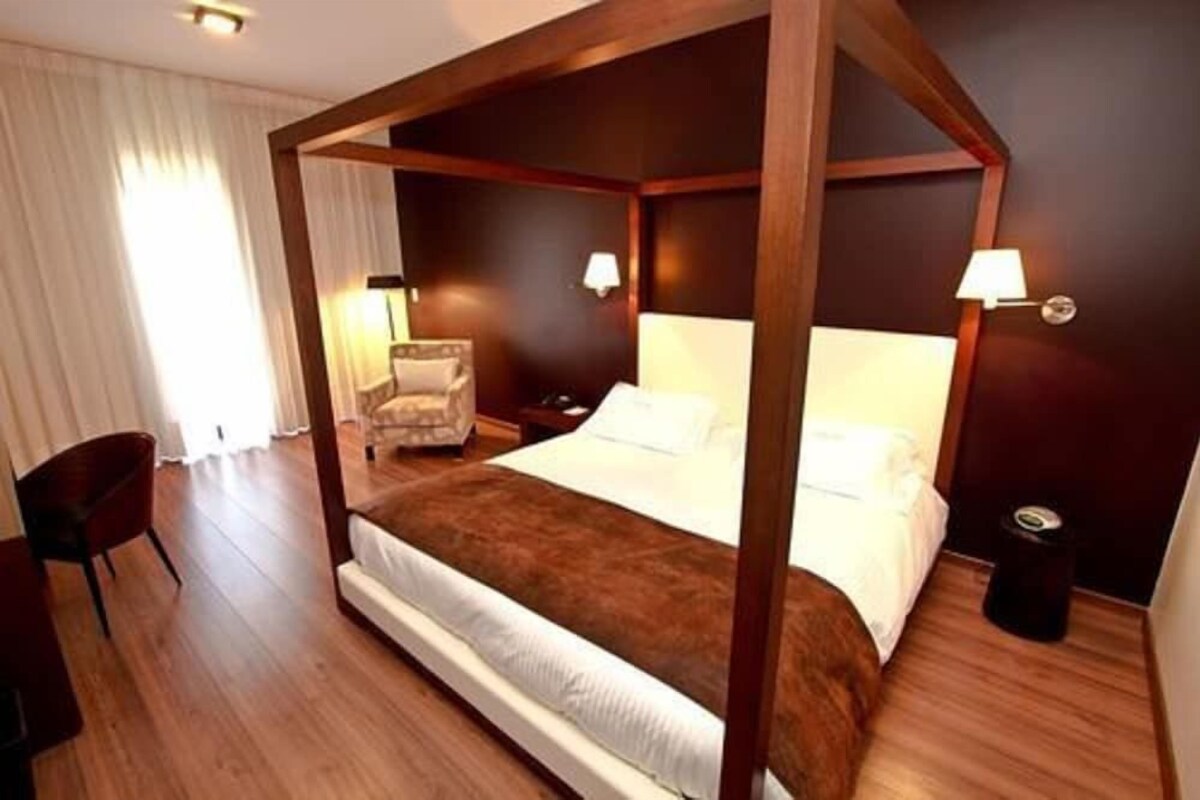 Una cama con dosel y estructura de madera, una silla, una mesita de noche y dos lámparas fijadas a la pared.