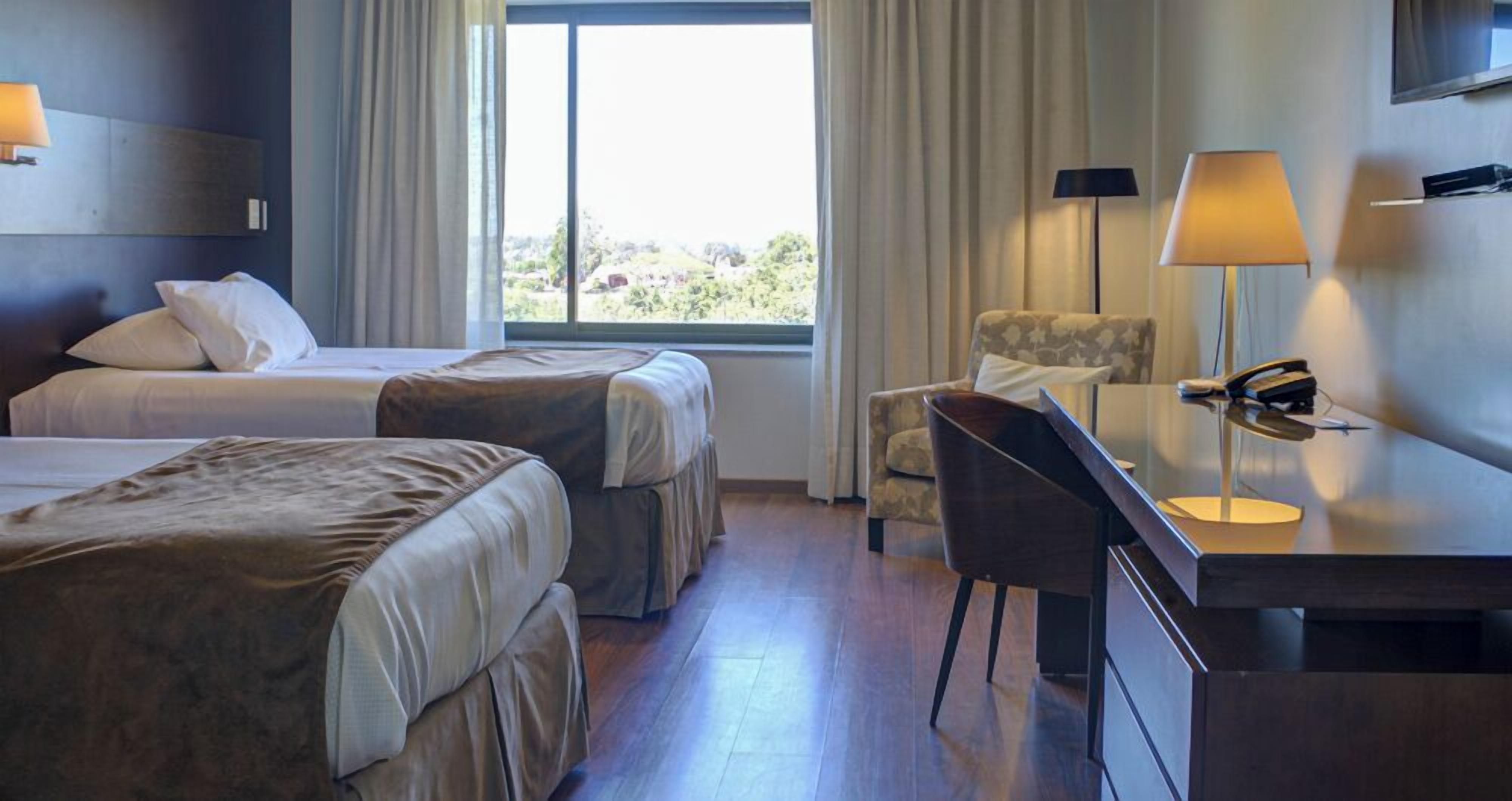 Una habitación de hotel con dos camas, un escritorio, una silla y un ventanal grande.