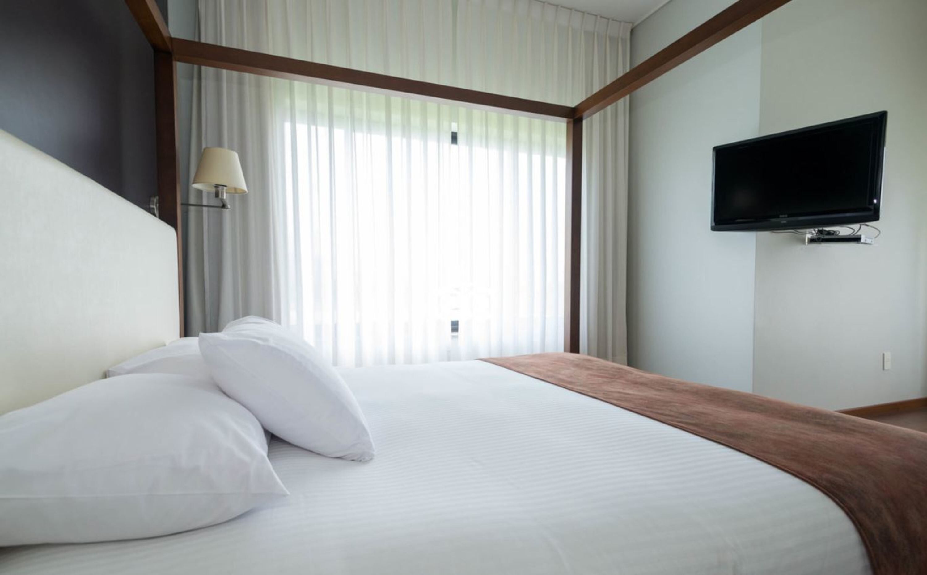 Una habitación de hotel con una cama grande, un televisor de pantalla plana montado en la pared y una ventana con cortinas translúcidas.
