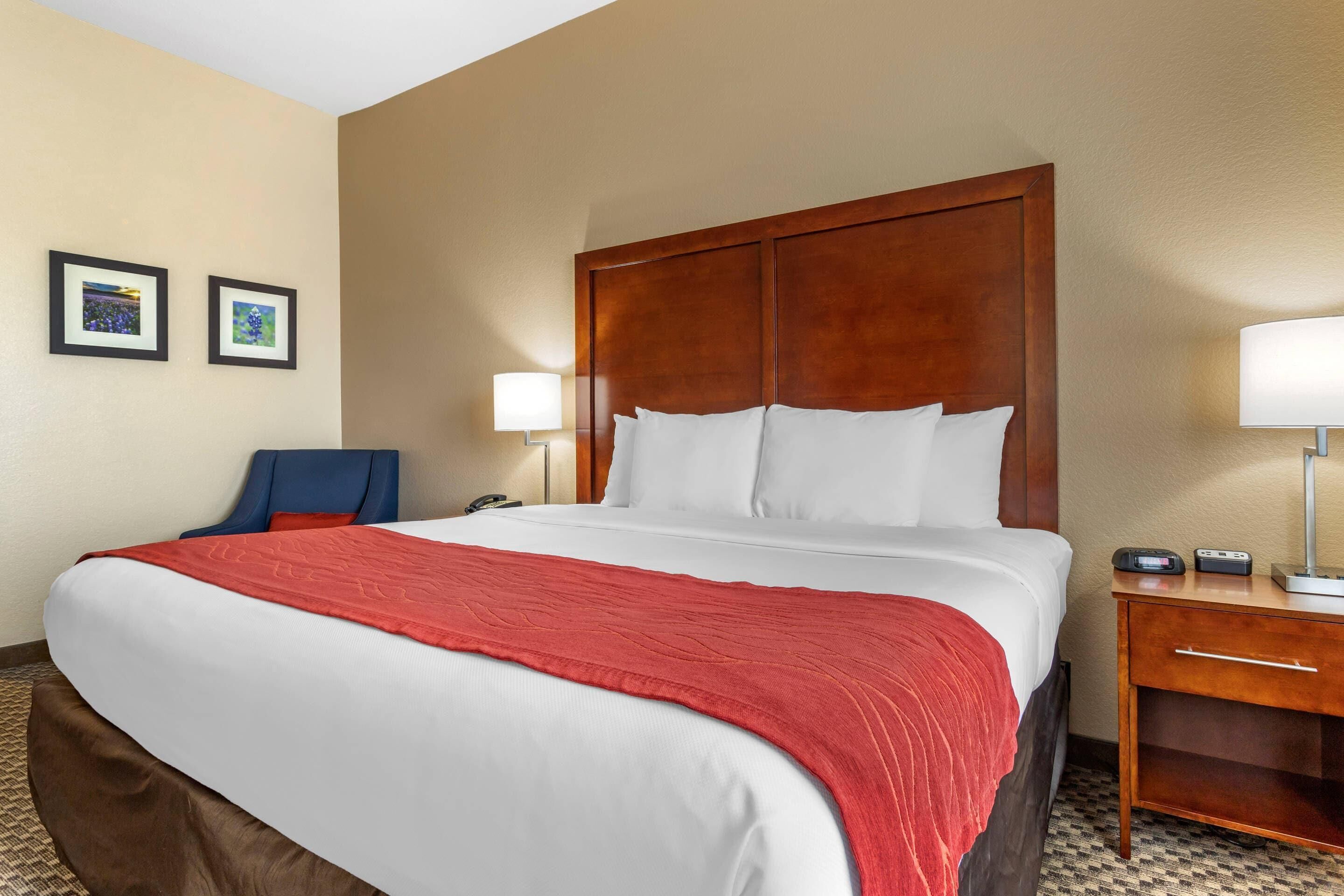 A hotel room with a large bed, a wooden headboard, two bedside lamps, a desk with a phone and a power strip, and two framed pictures on the wall.
