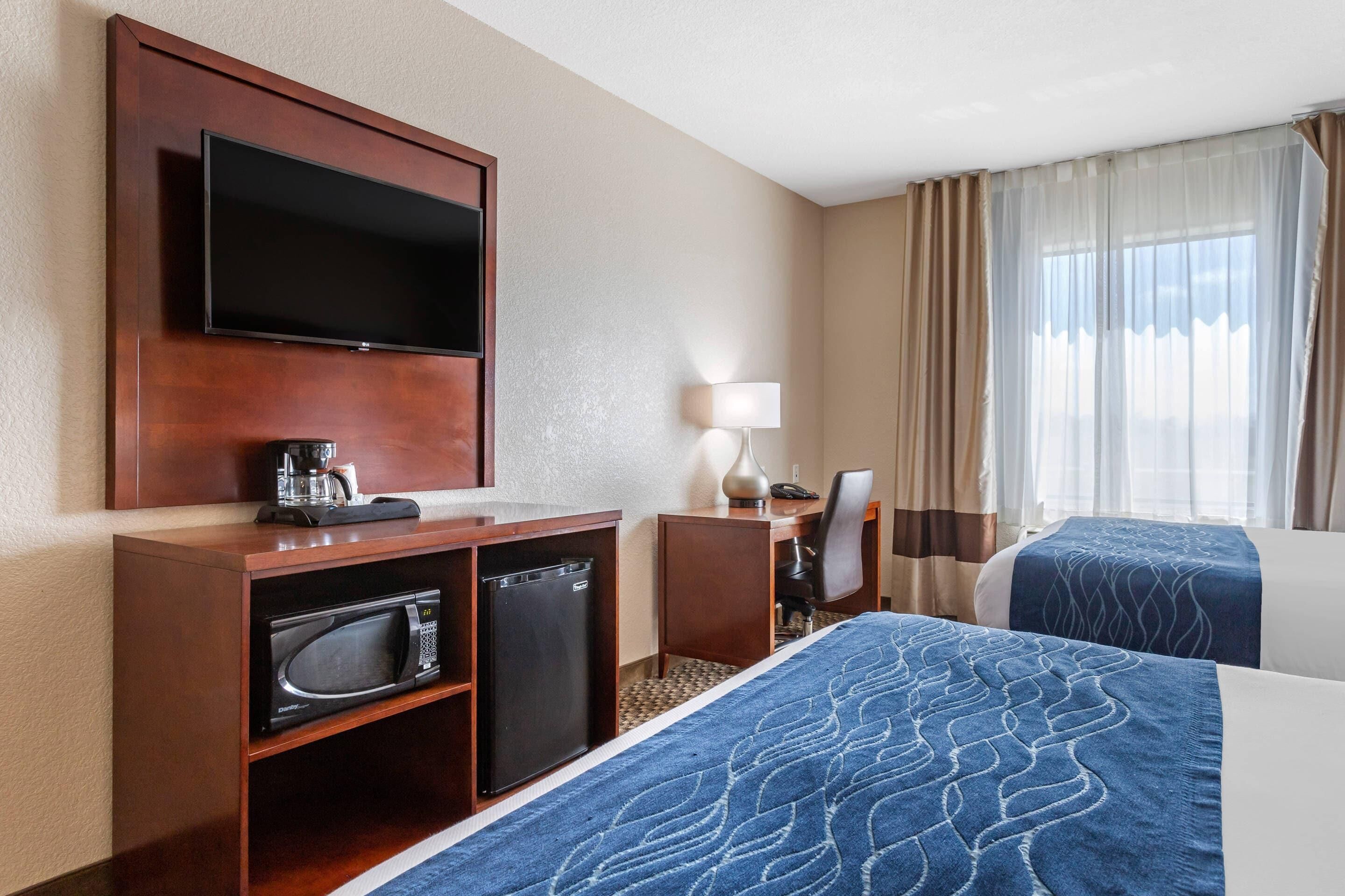 A hotel room with a flat-screen TV, a coffee maker, a microwave, a desk with a lamp, and two beds with blue bedding.