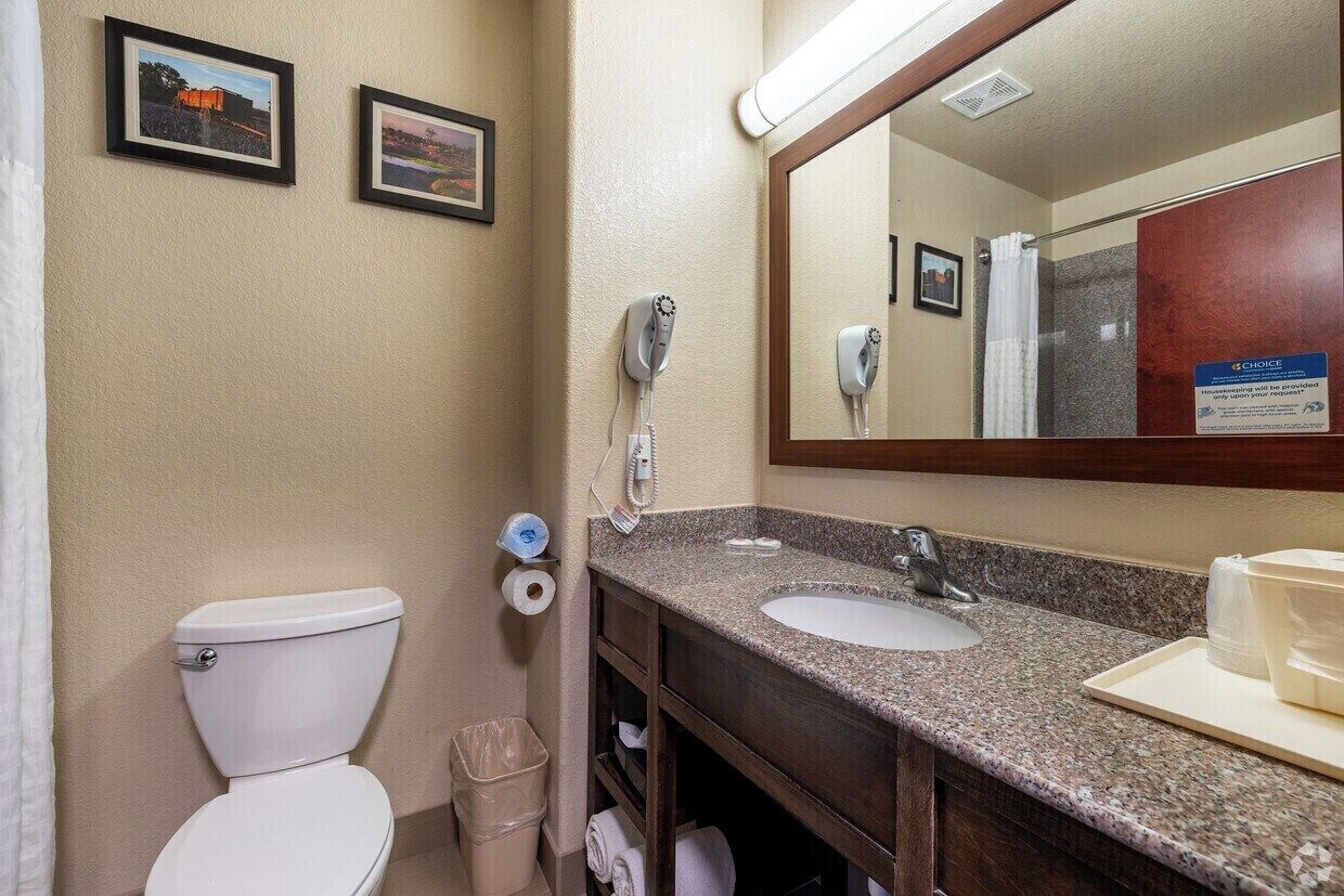 A bathroom with a toilet, sink, and mirror. There are two framed pictures on the wall, a hairdryer, and a trash can.
