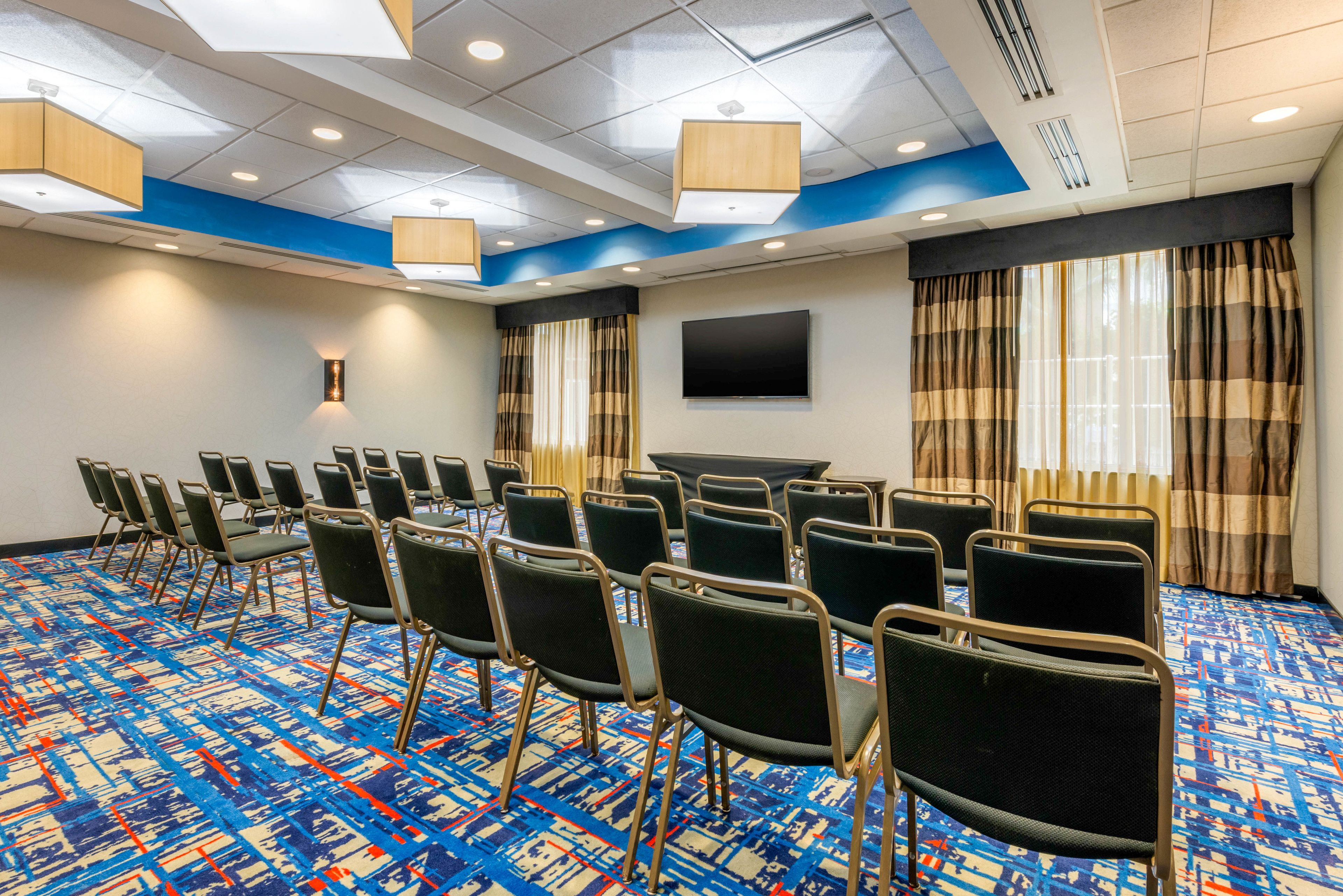 A conference room with a patterned carpet, rows of chairs, a flat-screen TV, and curtains.