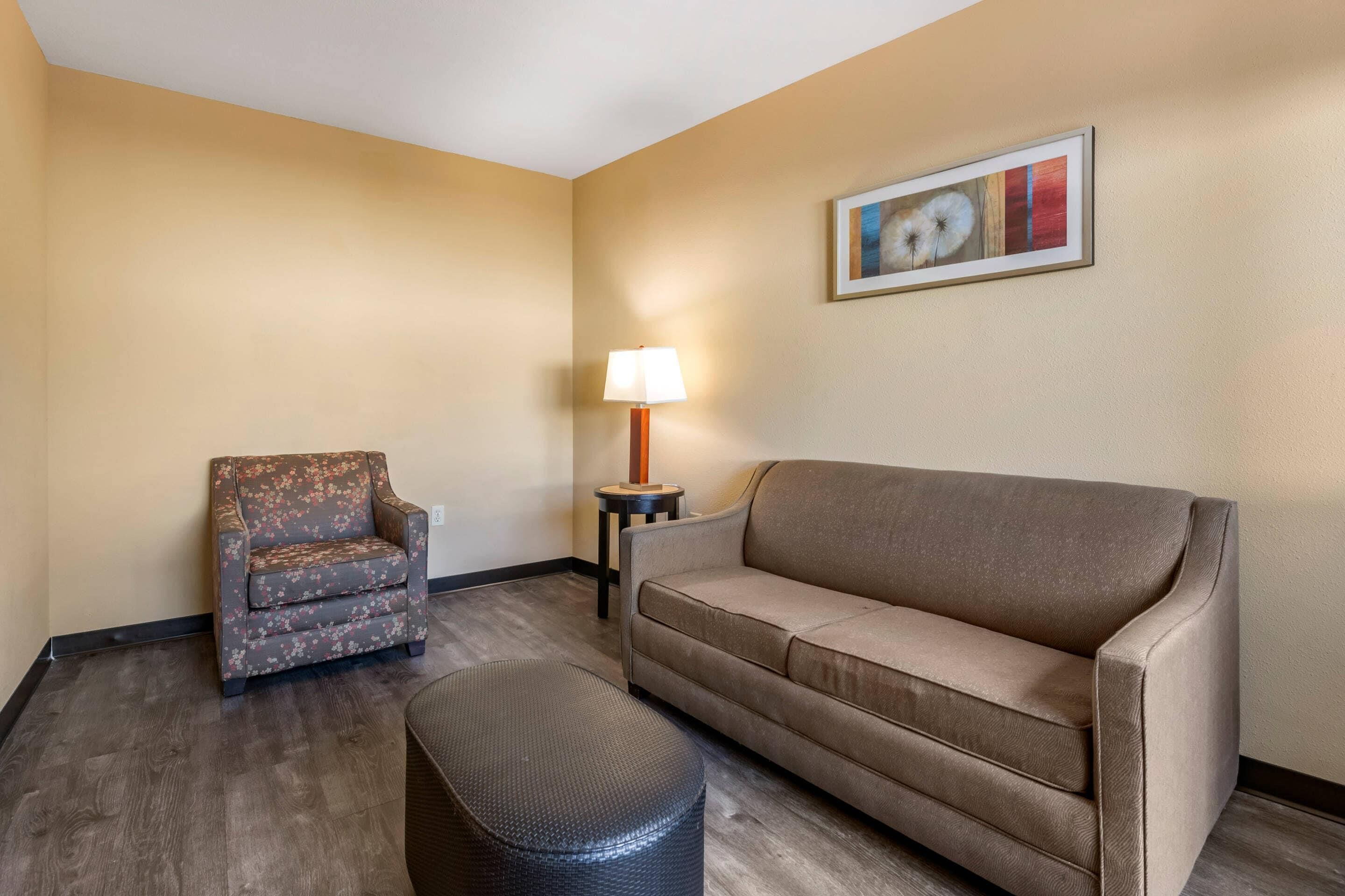Una habitación con un sofá color beige, un sillón floral, una mesita redonda con una lámpara y un cuadro en la pared.