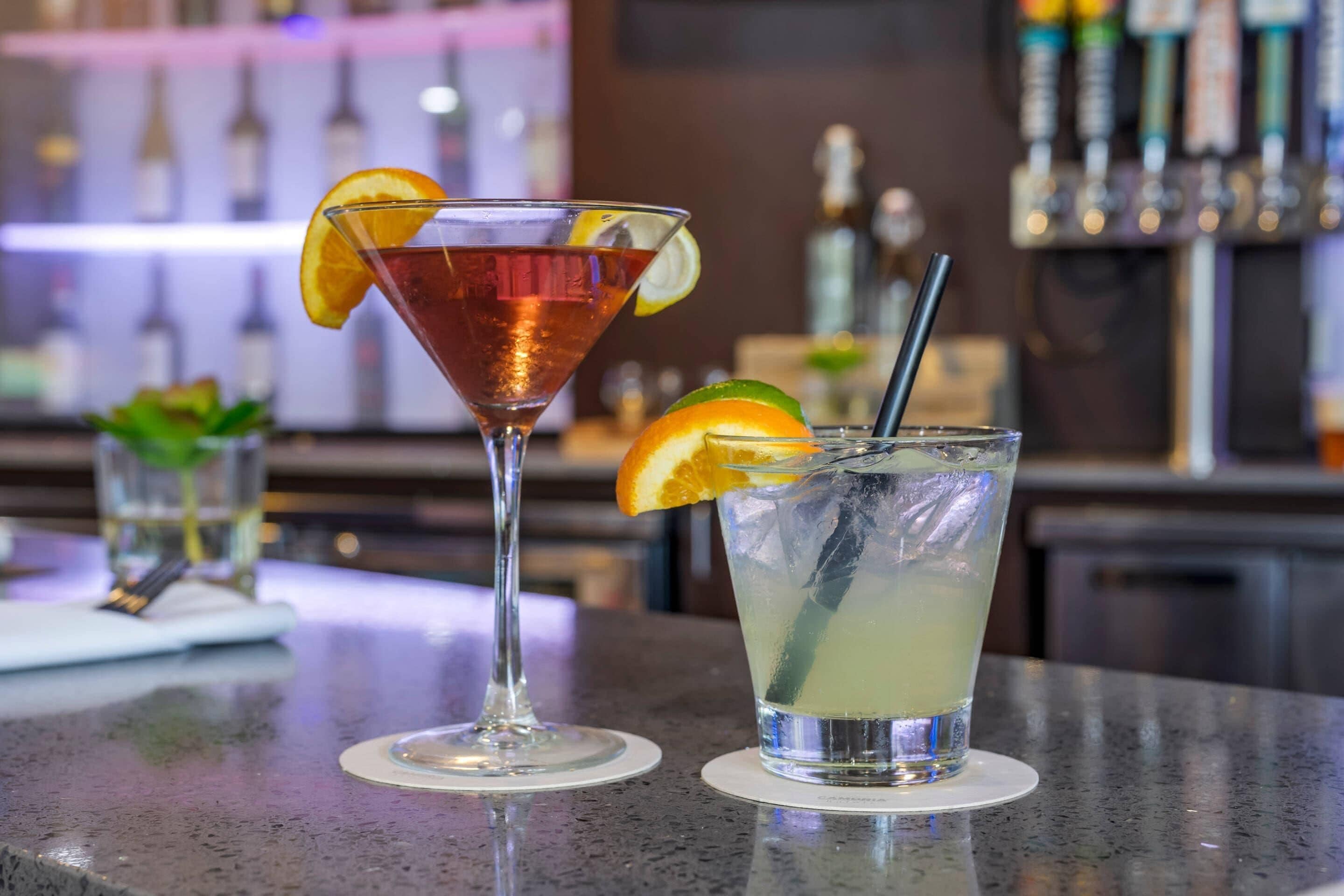 Dos bebidas sobre una barra, una en una copa de martini y otra en un vaso alto, con una barra y botellas al fondo.