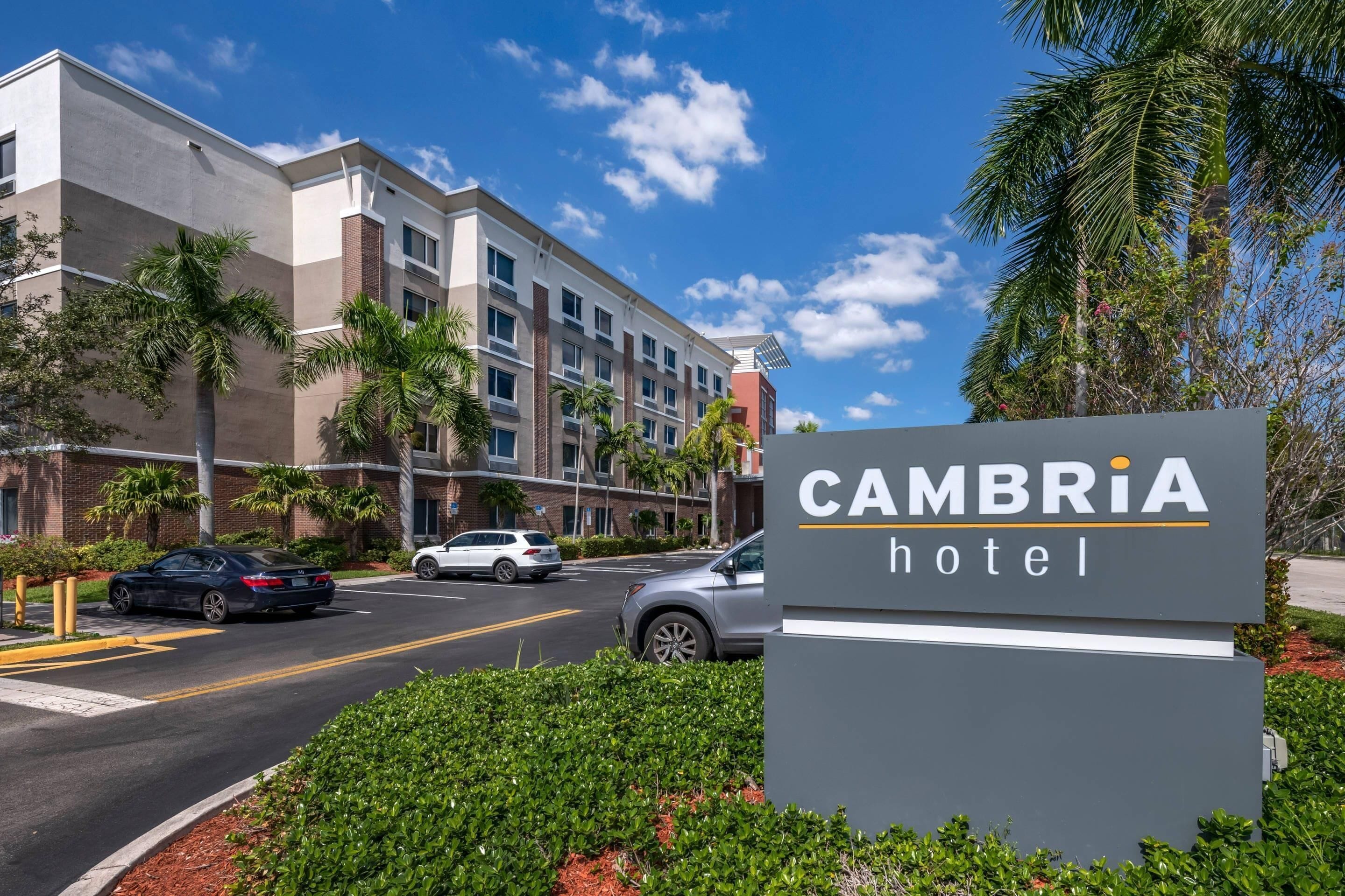 Un letrero de hotel con la inscripción 'CAMBRIA hotel', rodeado de follaje y palmeras.