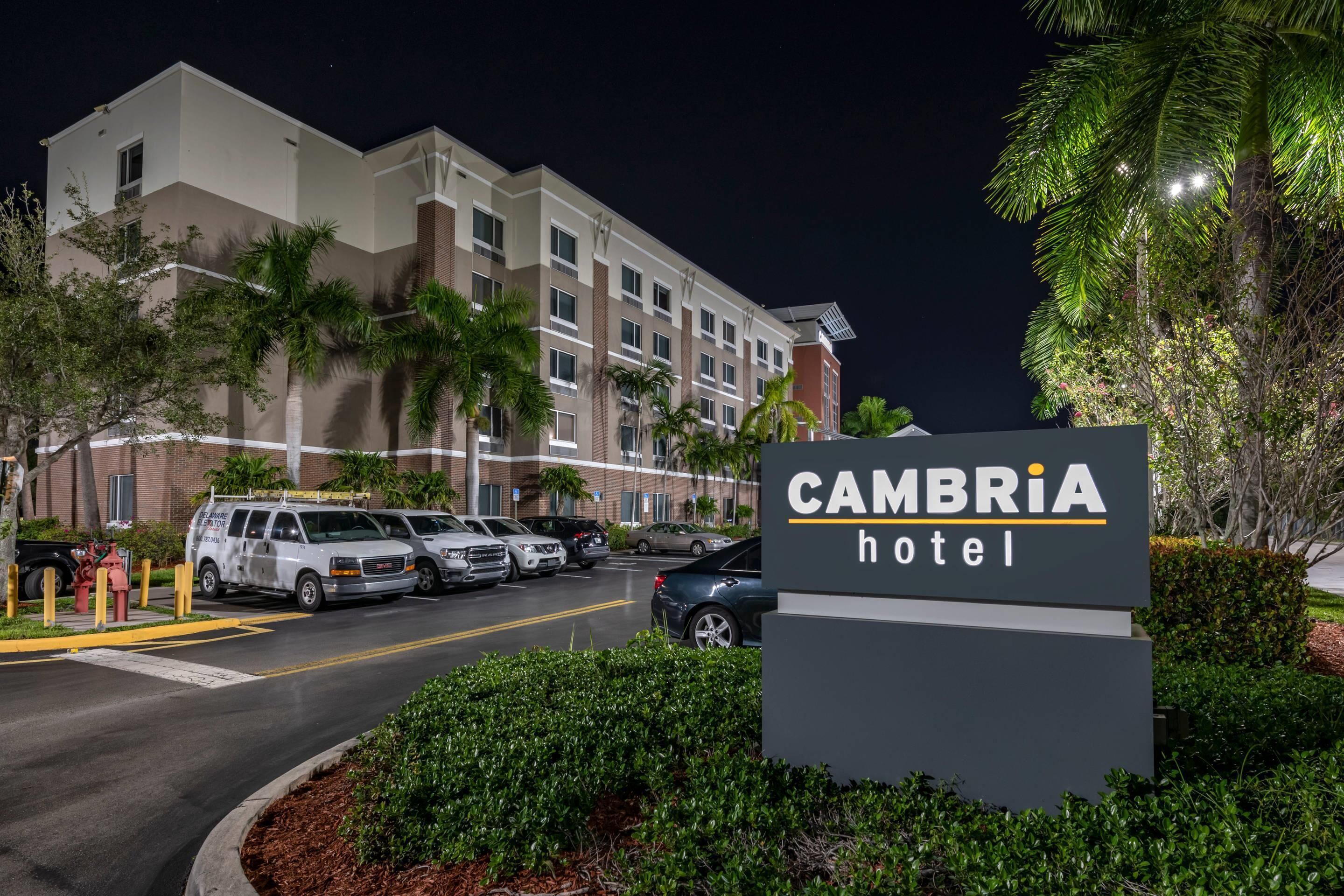 A hotel entrance with a sign reading "CAMBRIA hotel," a parking lot with several vehicles, and a building with multiple floors and windows.