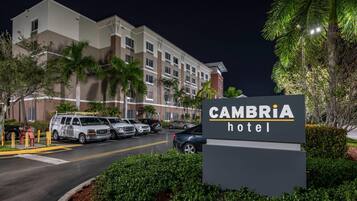 A hotel entrance with a sign reading "CAMBRIA hotel," a parking lot with several vehicles, and a building with multiple floors and windows.