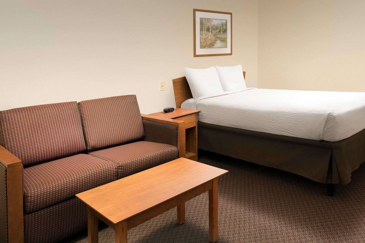 A hotel room with a brown patterned sofa, a wooden table, a bed with white bedding, and a framed picture on the wall.