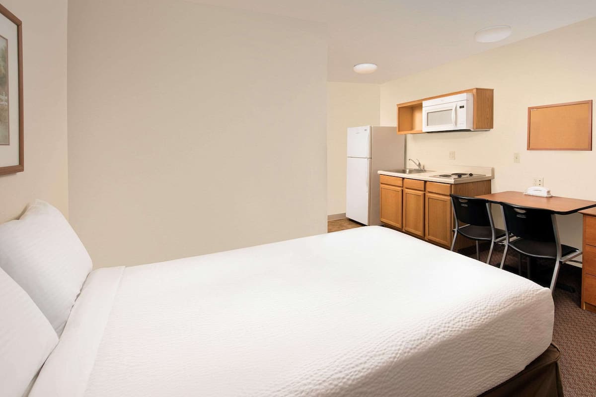 A hotel room with a bed, a desk, a microwave, a refrigerator, and a chair.