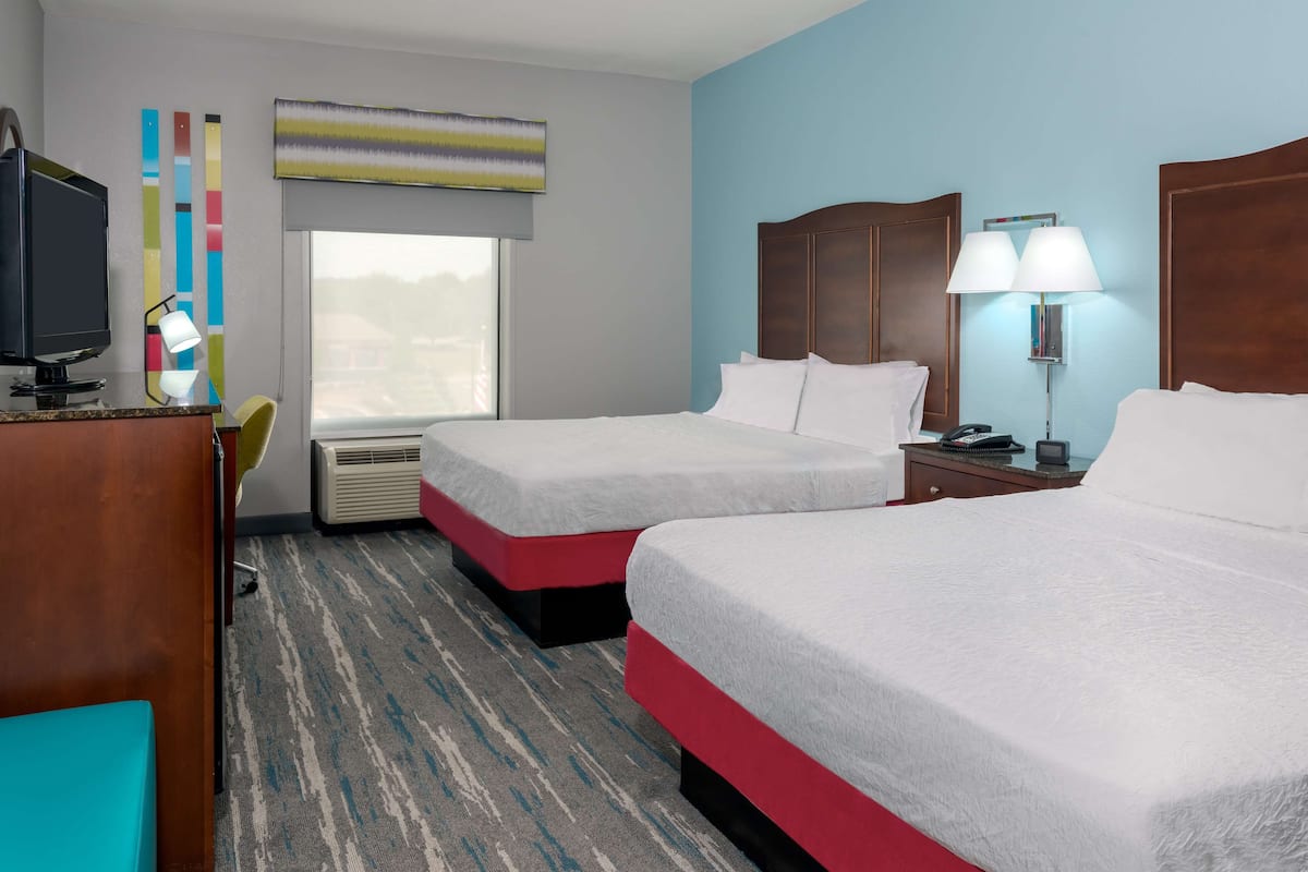 A hotel room with two beds, a desk with a computer, a TV, and a window with blinds.