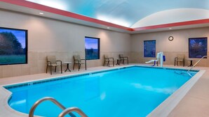 Una amplia piscina interior con agua cristalina y azul, rodeada de azulejos color beige y una pared de acento roja. Hay varias sillas y un reloj en la pared.