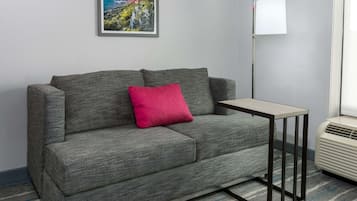 A grey sofa with a red pillow, a small table, a lamp, and an air conditioner in a room with a carpeted floor.