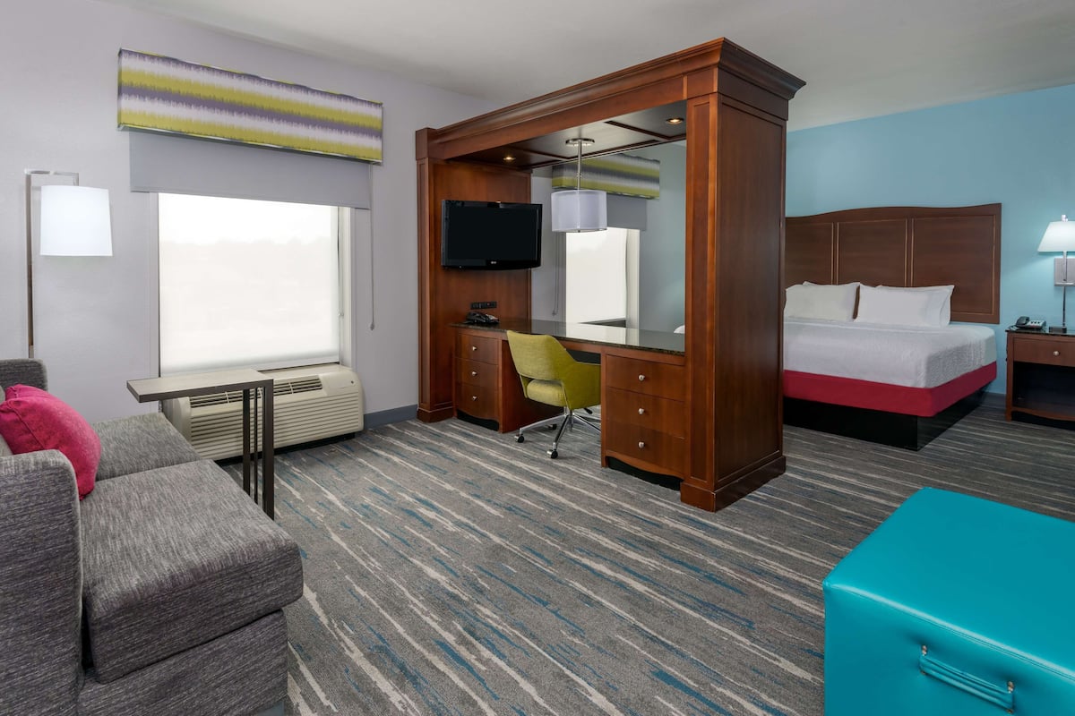 A hotel room with a bed, a desk with a chair, a television, a window, and a large mirror.
