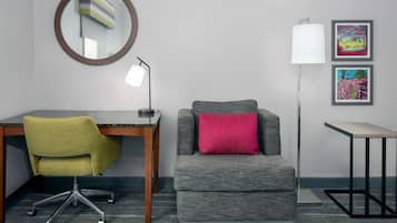A hotel room with a desk, office chair, grey armchair, lamp, and two framed pictures on the wall.