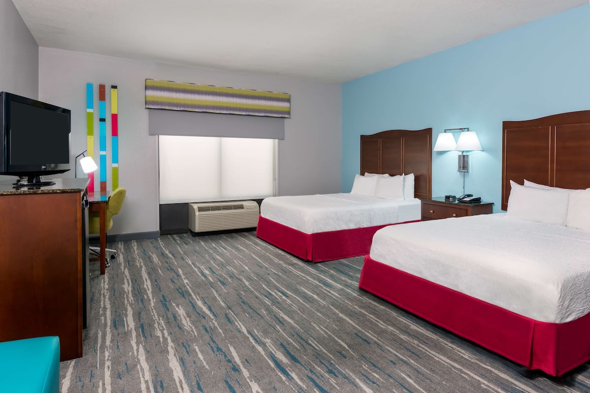 A hotel room with two beds, a television, a desk, a chair, a window, and a wall-mounted air conditioner.