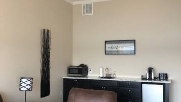 A living room with a brown leather sofa, a coffee table with a remote control, a wall-mounted lamp, a kitchen area with a microwave, a coffee maker, and a framed quote on the wall.