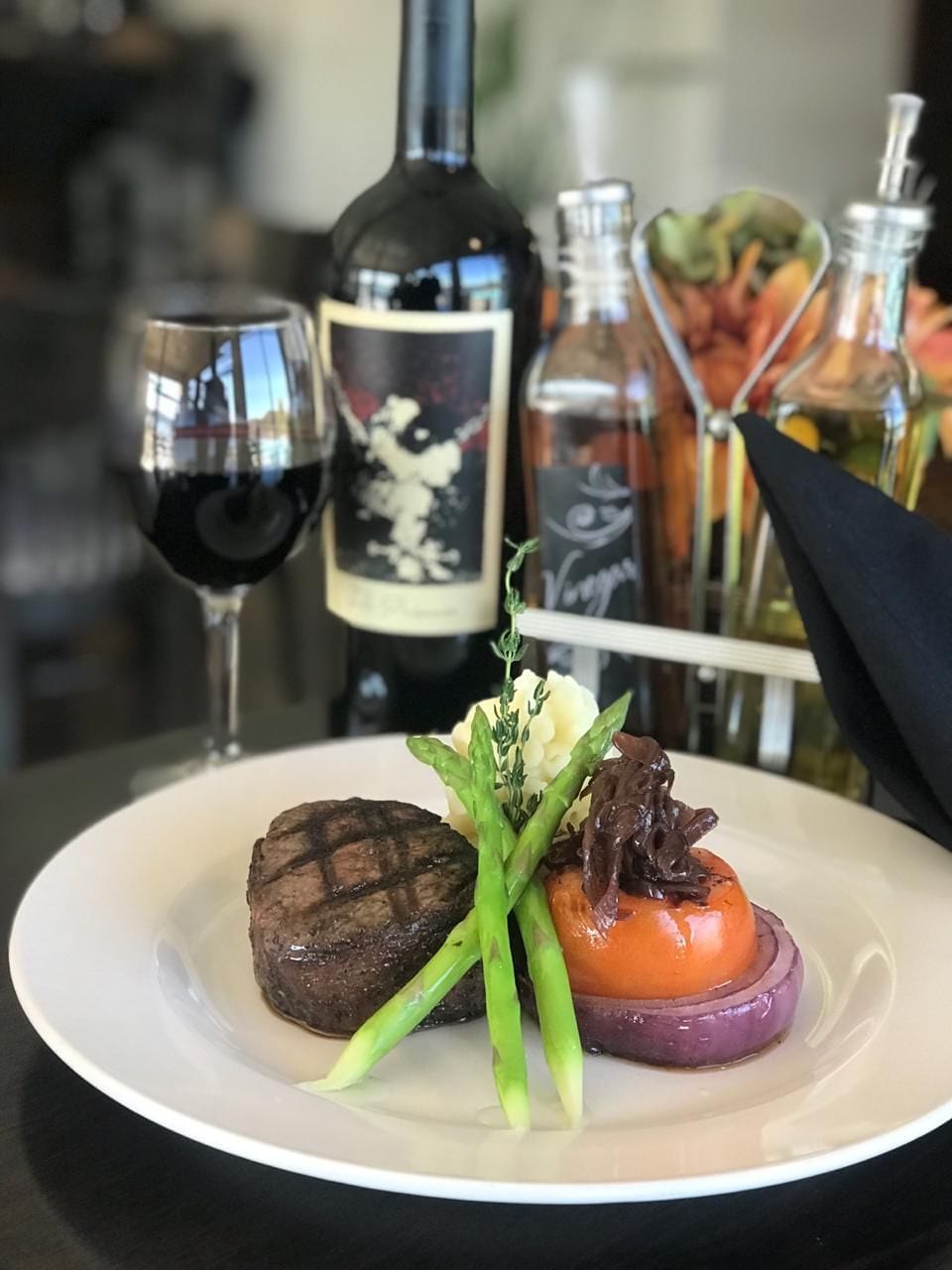 A plate of food with a steak and vegetables, a wine glass, and bottles of wine on a table.