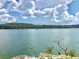 A lake with a boat, rocky shoreline, and a forested hill in the background.