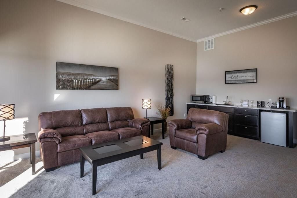 A living room with a brown leather sofa, a coffee table, a side table with a lamp, a decorative vase, and a kitchen area with a refrigerator and microwave.