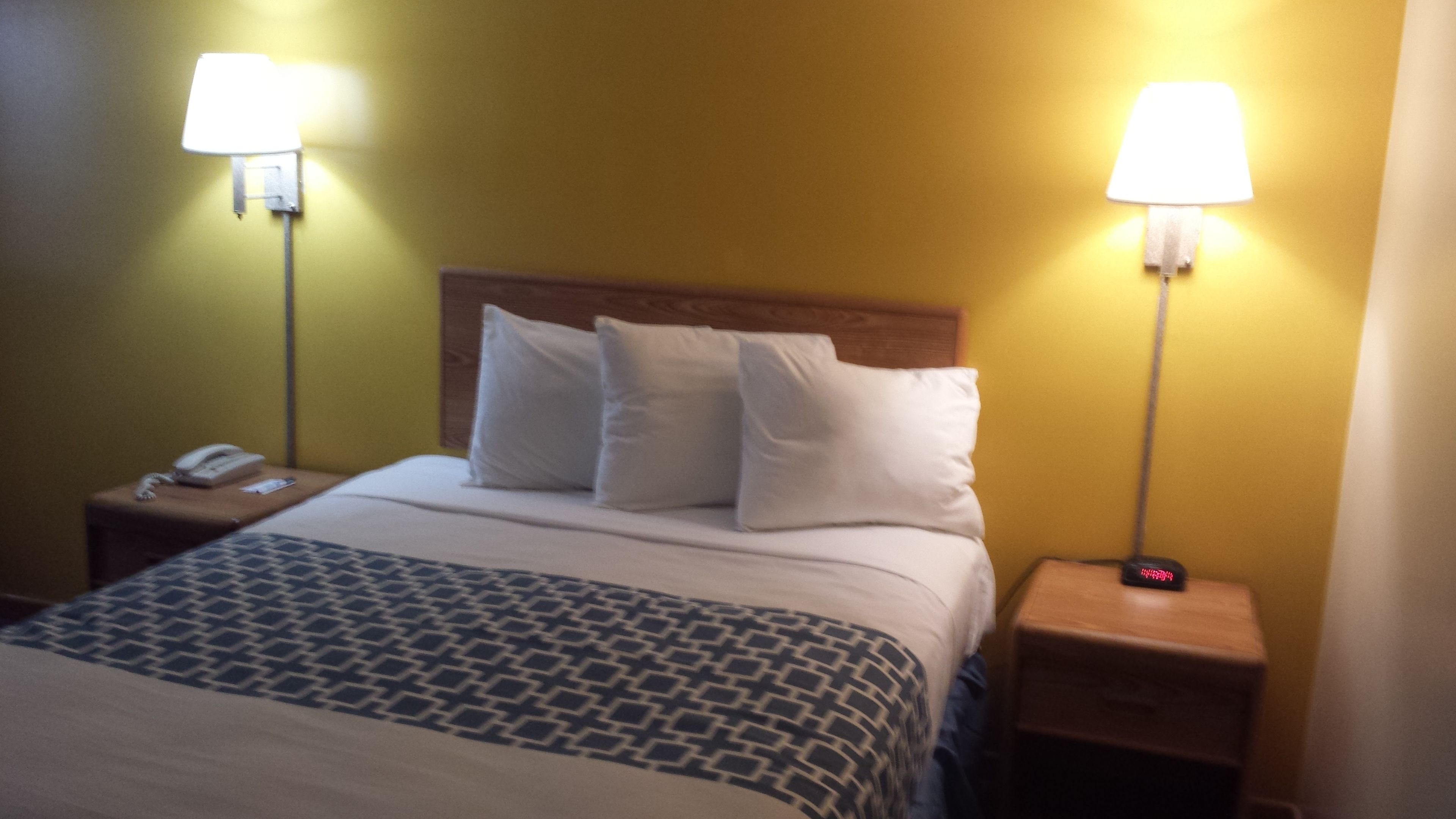 A hotel room with a bed, two bedside lamps, a nightstand with a phone, and a digital clock.