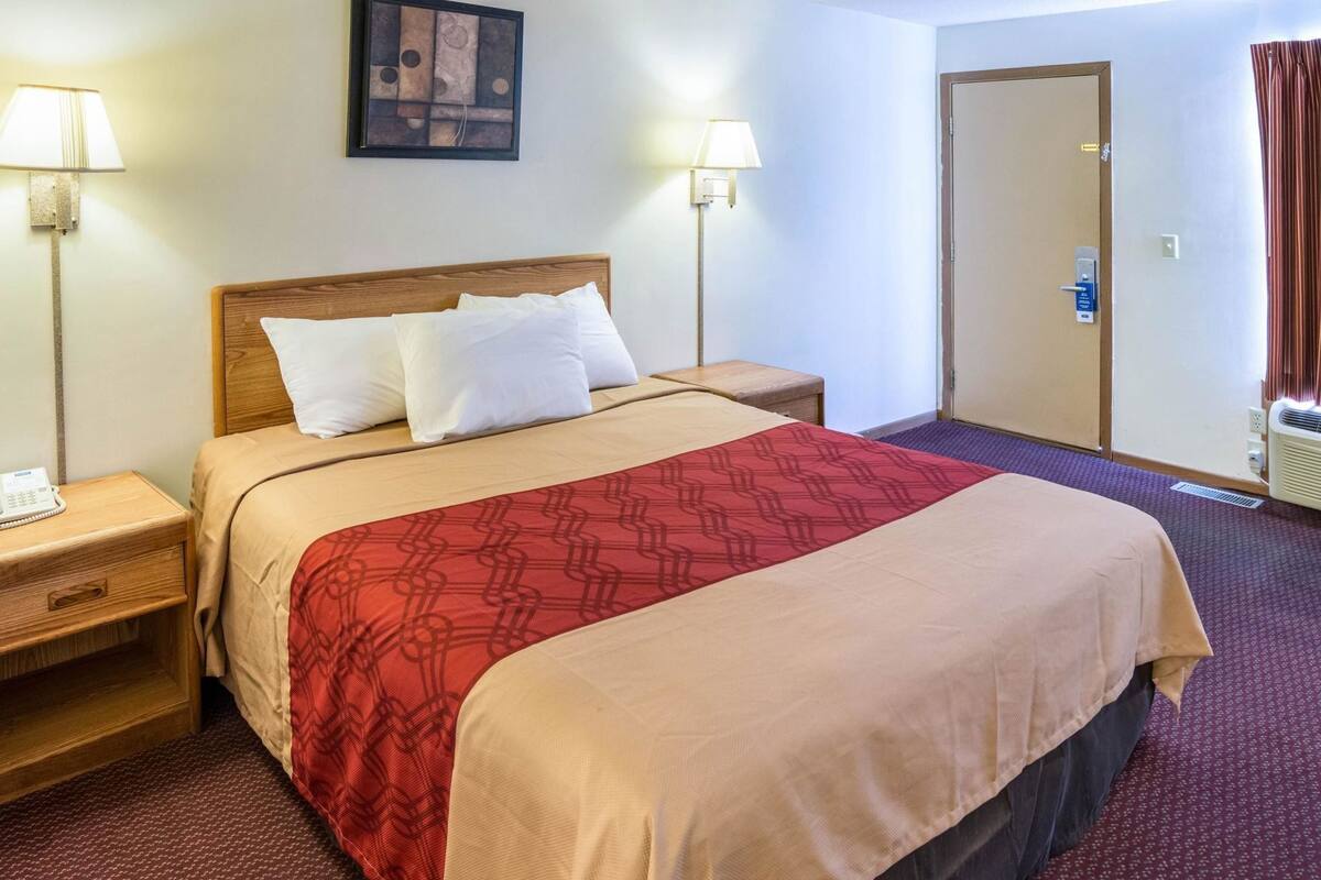 A hotel room with a bed, two bedside lamps, a nightstand, a door, and a wall-mounted artwork.